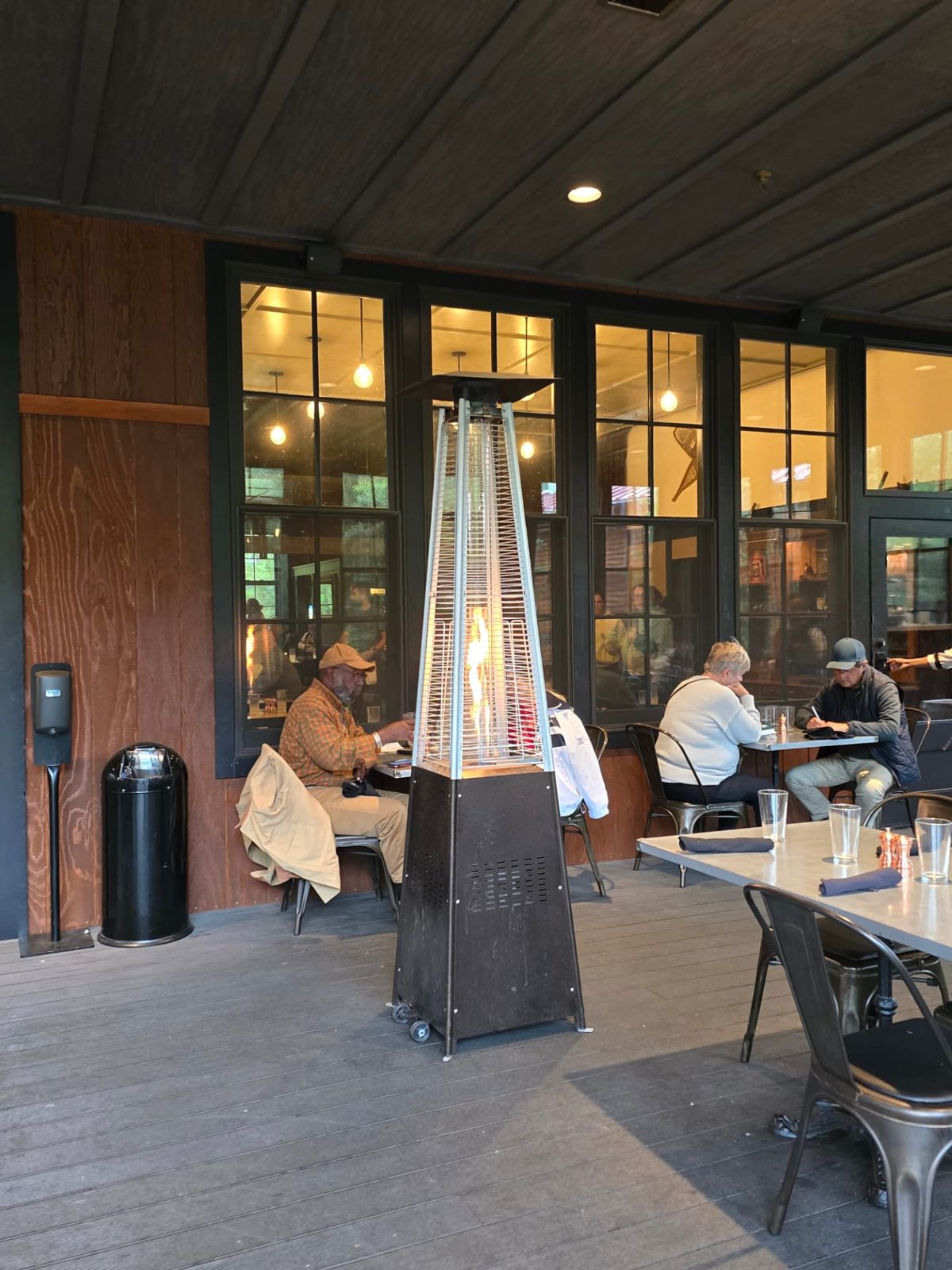 Covered wooden patio with a tall glowing pyramid patio heater, travelers seated at small metal tables in front of warmly lit windows