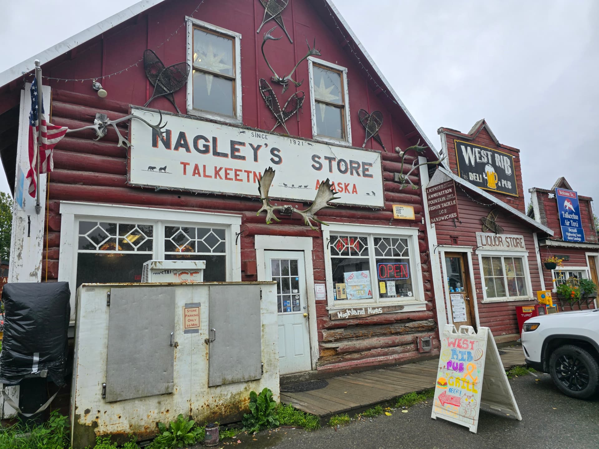 Red log-cabin storefront of Nagley’s Store in Talkeetna, Alaska, with moose antlers mounted over the front door, snowshoes and antlers on the siding, an American flag at the corner, and the West Rib Deli & Pub beside it on a damp, gray day